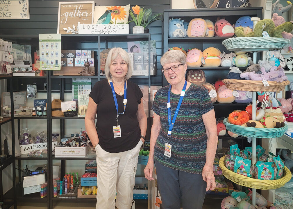 Two volunteers standing beside each other in front of a display wall inside the gift shop at Markham Stouffville Hospital.
