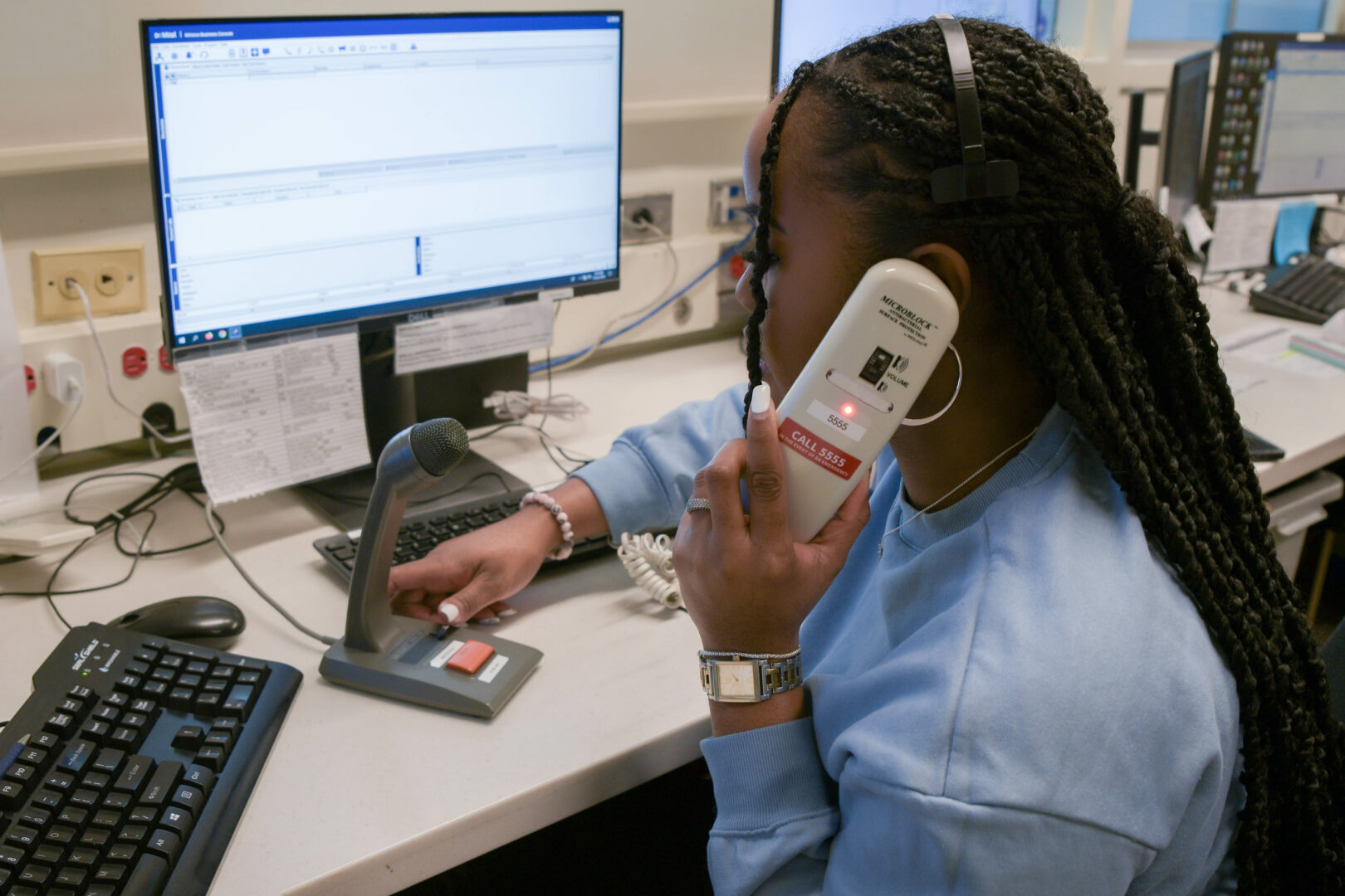 Image of the communications clerk, Herbeisha Williams, conducting code calling at the markham stouffville hospital in her office, holding a phone and a microphone on the other hand.